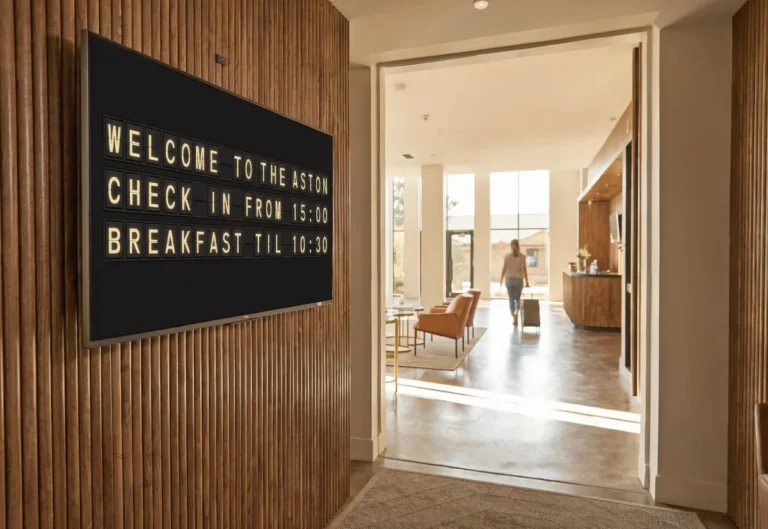 A guest arriving in a hotel lobby with a television showing a split-flap style welcome board with check in and breakfast times.