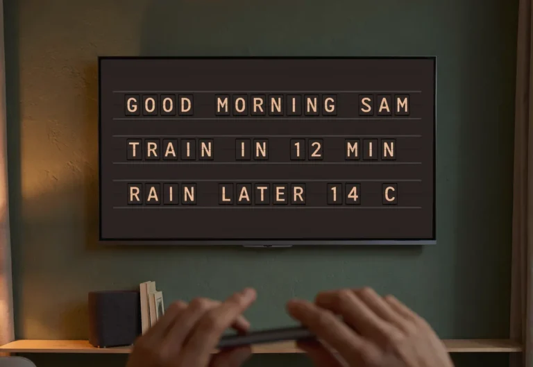 A phone held in the foreground of a living room with a television showing a split-flap style morning board.