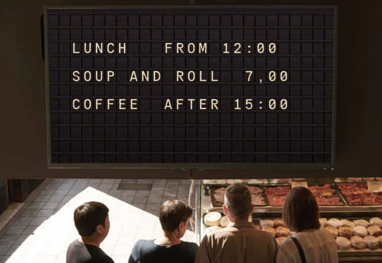 A queue at a deli counter beneath a television showing a split-flap style board of lunchtime service times.