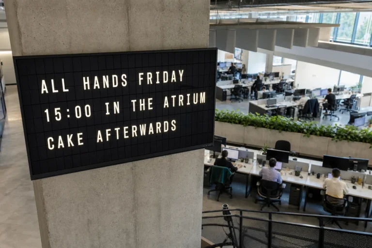 Looking down over an open plan office at a pillar mounted television showing a split-flap style all hands announcement.