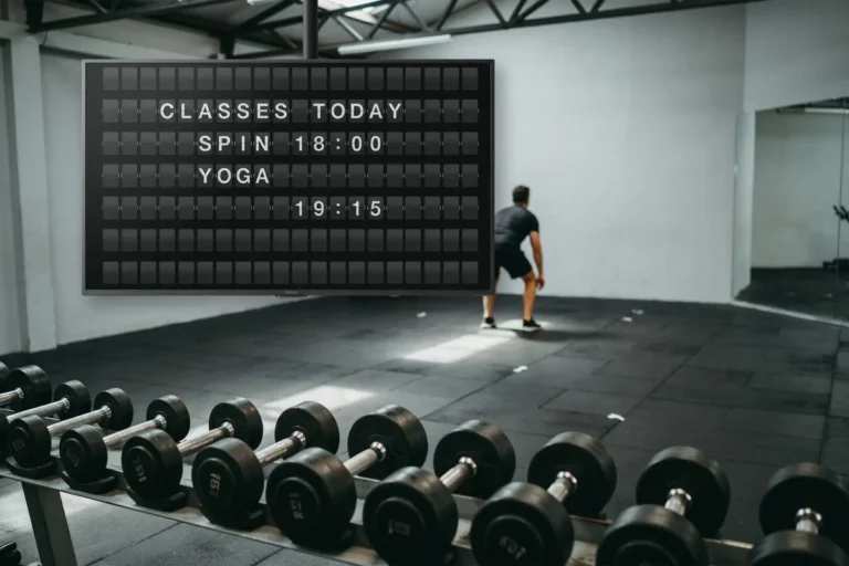 A gym floor with a television high on the wall showing a split-flap style board of today's class times.